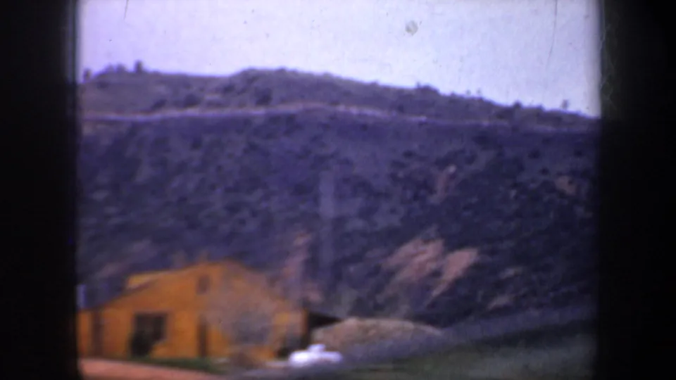 1957 Oregon home-movie frame showing a small yellow building below a steep hillside and distant ridge, with soft 8mm color shift.