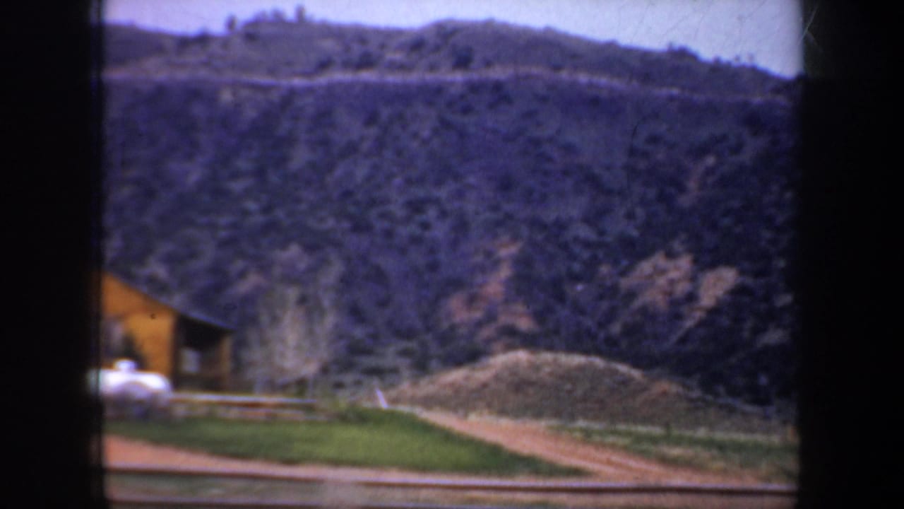 1957 Portland, Oregon home-movie frame showing a tree-dotted hillside, a small yellow structure, and a pale object near the roadside foreground.