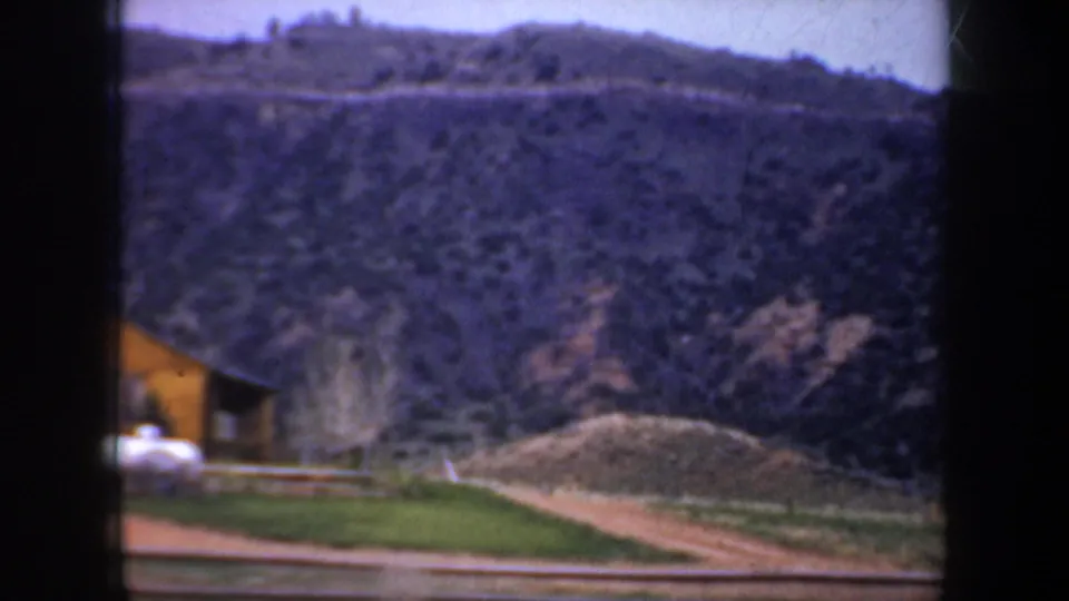 1957 Portland, Oregon home-movie frame showing a tree-dotted hillside, a small yellow structure, and a pale object near the roadside foreground.