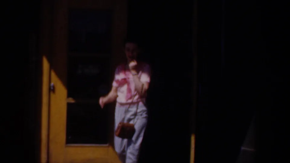 1957 8mm home-movie frame showing a woman in a pink top and light pants waving from a doorway while holding a small purse.