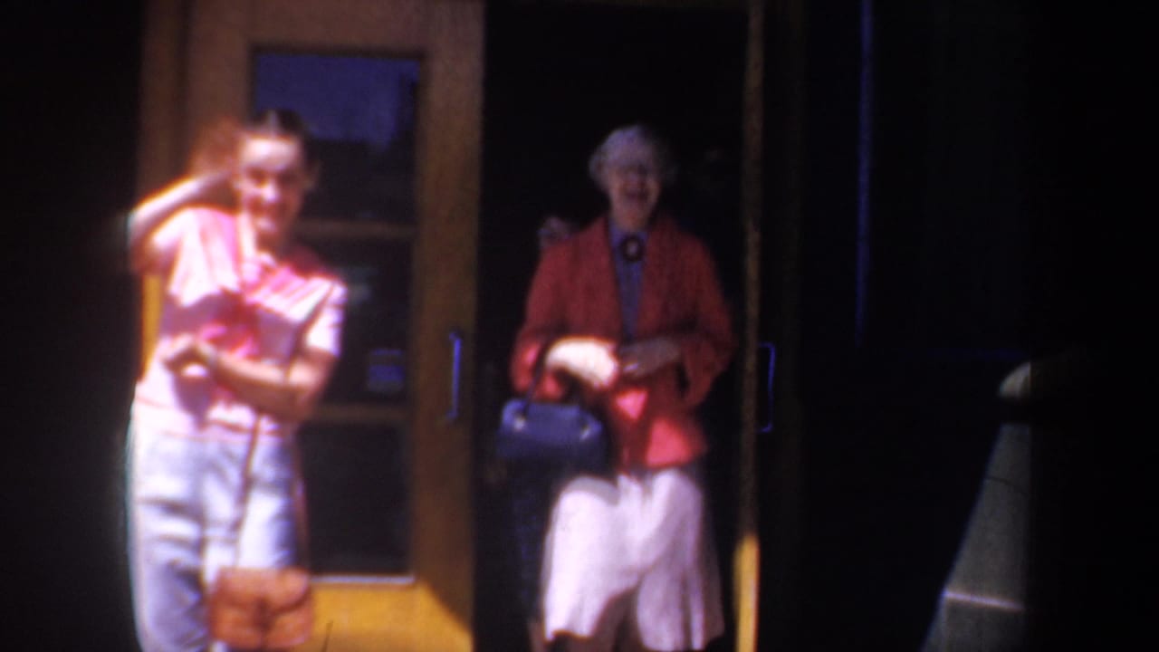 Woman in pink waves near a doorway while an older woman in a red jacket stands nearby.