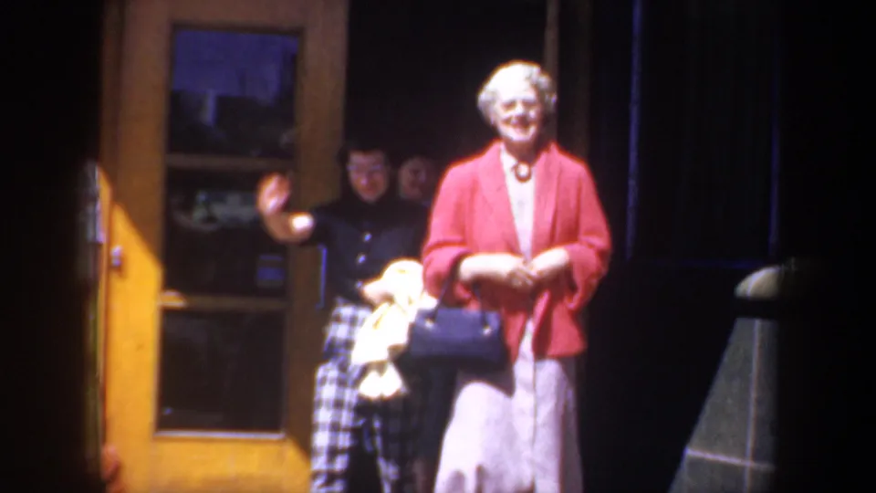 An older woman in a red jacket and light skirt smiles near a yellow doorway, with other figures partly visible behind her.