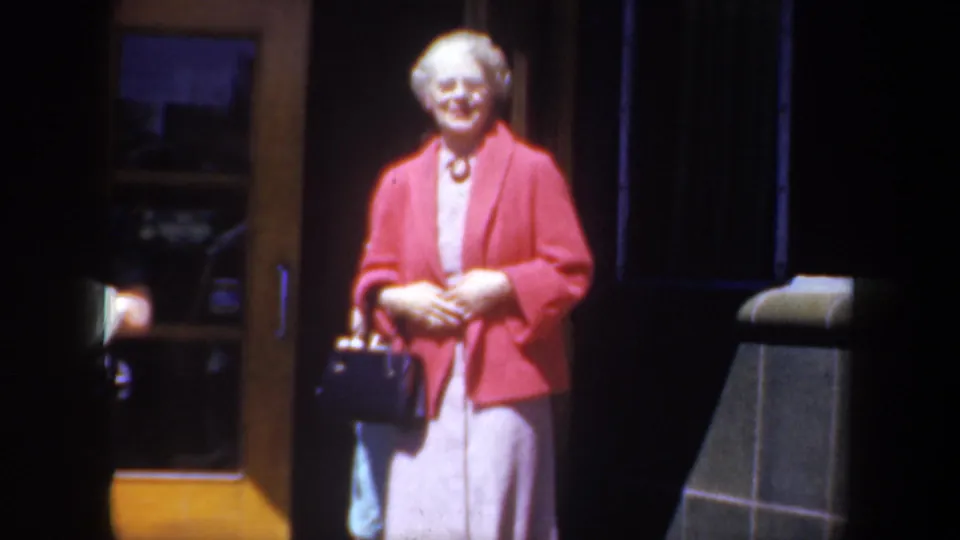 The older woman remains by the building entrance, smiling with a dark handbag in warm 1957 Portland home-movie footage.