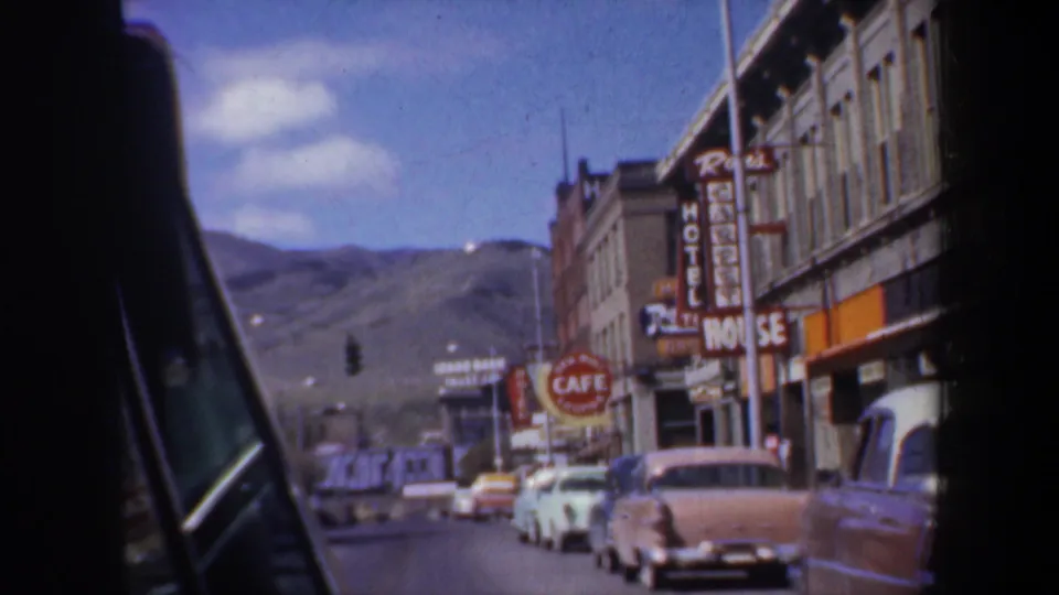 A passenger-view street scene in 1957 Oregon shows classic cars, commercial signs, storefronts, and distant hills under blue sky.
