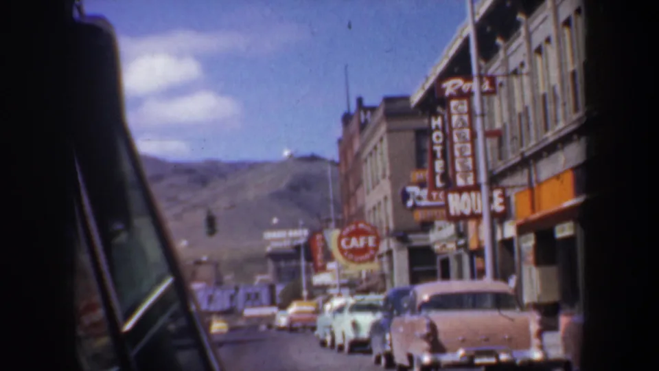 A 1957 Oregon street view from a moving car shows vintage automobiles, cafe and hotel signage, storefront facades, and hills in the background.