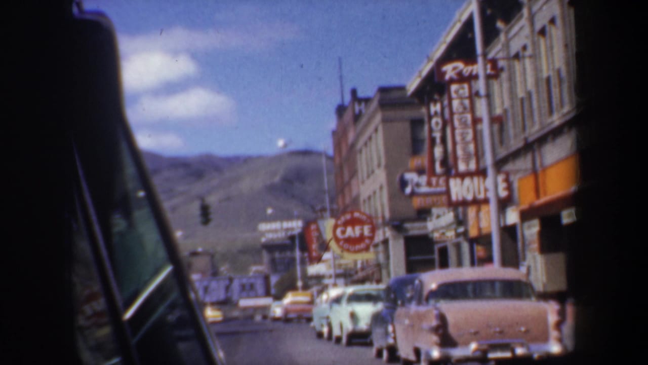 1957 Portland, Oregon street view from a moving car, with parked mid-century automobiles, hotel and cafe signs, storefronts, and distant hills.