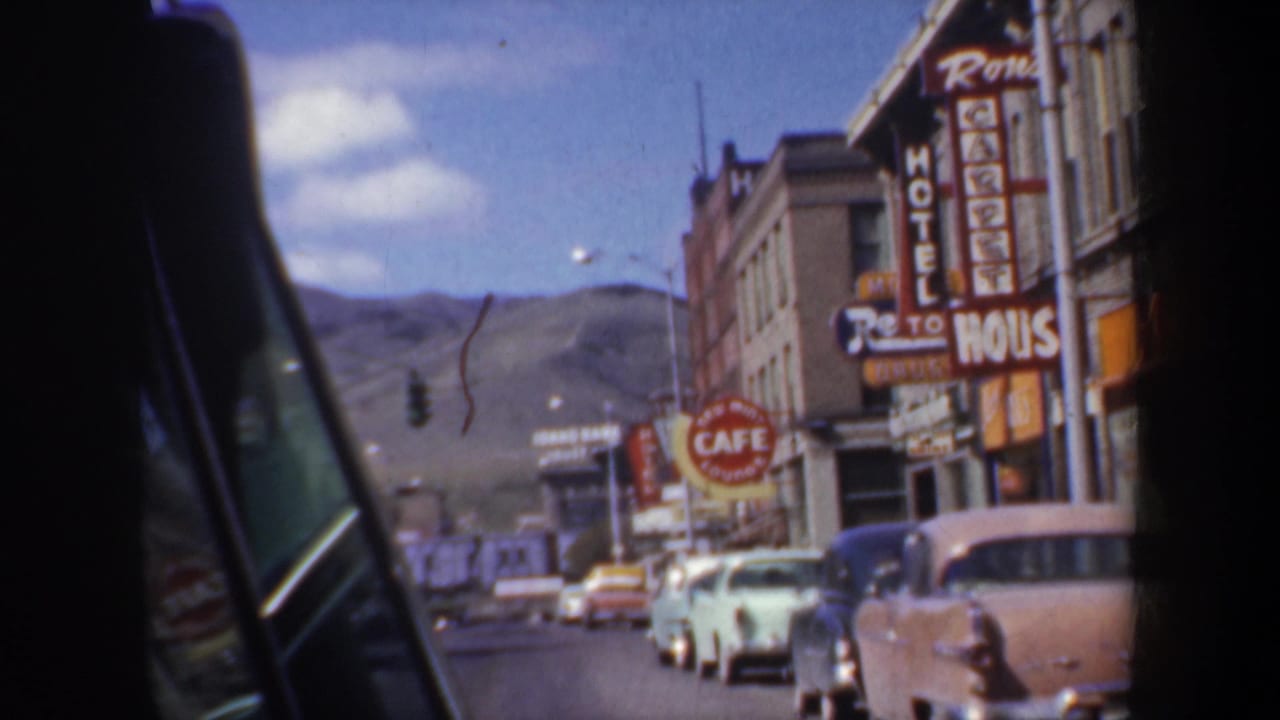 Streetfront with hotel and cafe signs, parked cars, older buildings, and hills beyond the road.
