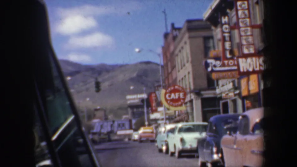 1957 Portland street footage from inside or beside a car, with neon business signs, parked automobiles, storefronts, and hills in the distance.
