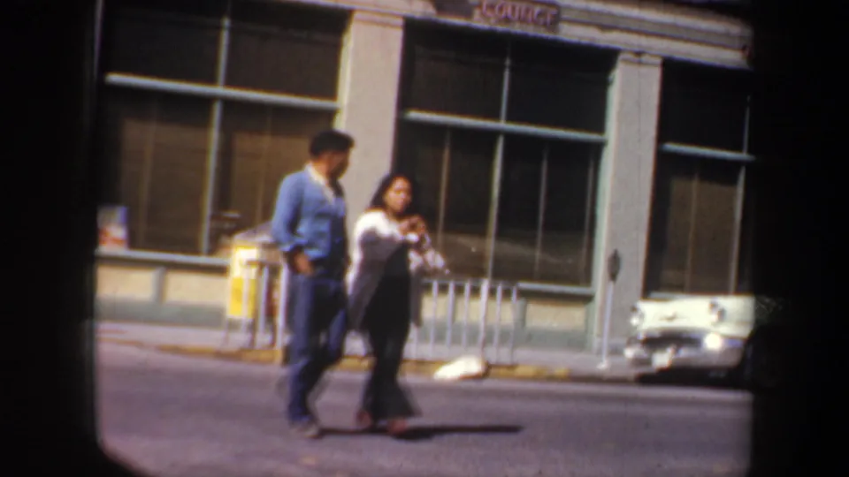 Two people walk along a Portland sidewalk in the 1957 home movie, passing storefront windows and curbside street fixtures.