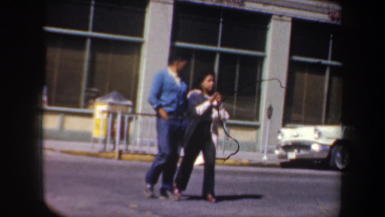 Two adults walk along a Portland street in supplied 1957 8mm footage, near storefront windows and a parked white car.