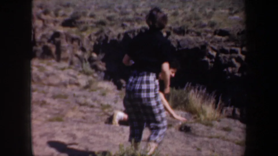 1957 home-movie frame of two travelers in rocky arid terrain, with one adult in plaid pants standing and another crouched low nearby.