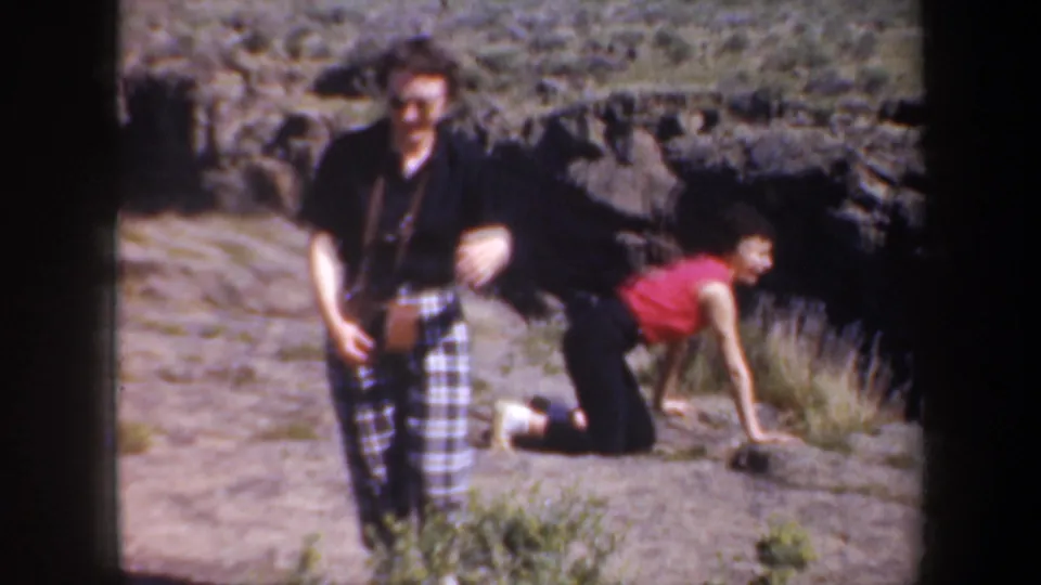 1957 8mm roadside scene with an adult facing the camera and another traveler in a red top crawling beside rocks and scrub.