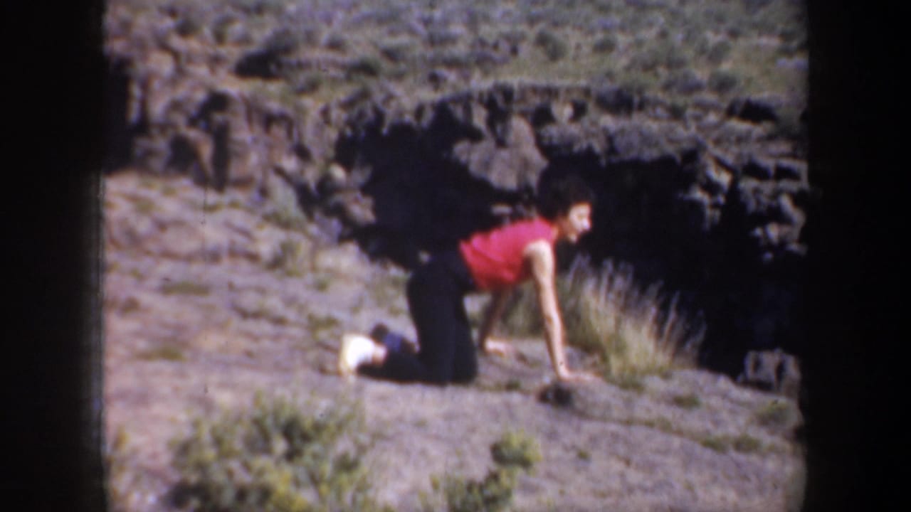 1957 Oregon home-movie frame showing a woman in red crawling over uneven rocky terrain with dark canyon rocks behind her.