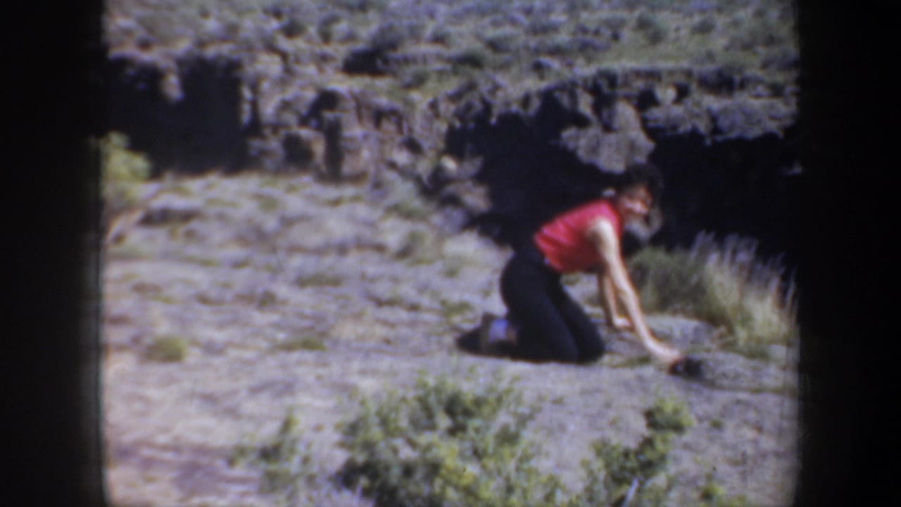 1957 Oregon home-movie frame of a woman in a red sleeveless top kneeling on rough canyon terrain, partly turned toward the ground.