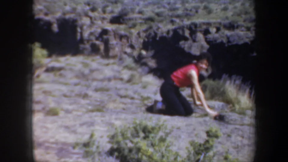 1957 Oregon home-movie frame of a woman in a red sleeveless top kneeling on rough canyon terrain, partly turned toward the ground.