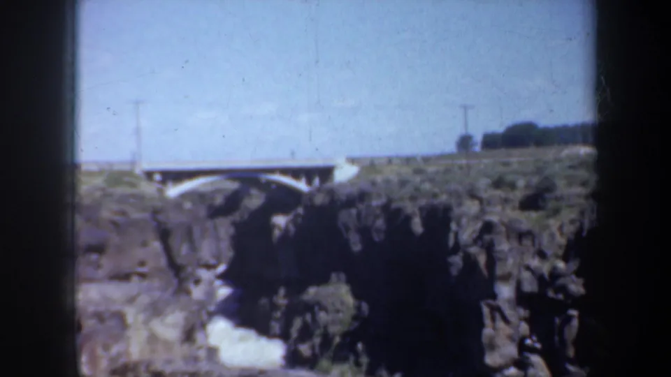 1957 Oregon home-movie frame showing a white arched bridge crossing a rocky canyon with river water visible below.