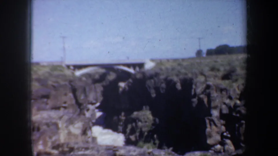 1957 Oregon home-movie frame with a white arched bridge, dark canyon cliffs, pale river water, and open sky.