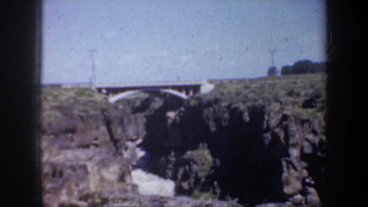 1957 Portland-area 8mm frame showing a pale arched bridge spanning rugged cliffs with scrub vegetation and utility poles.