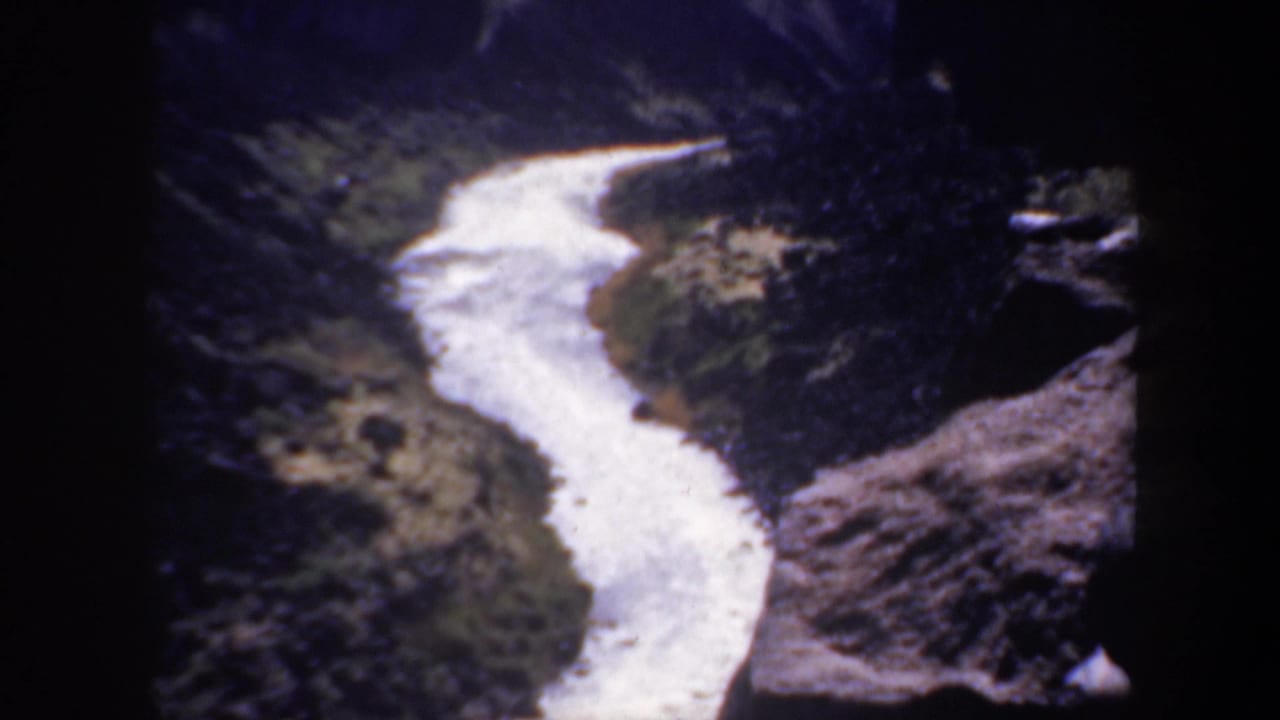 1957 Oregon home-movie frame showing a pale winding watercourse between dark canyon walls and sparse vegetation.