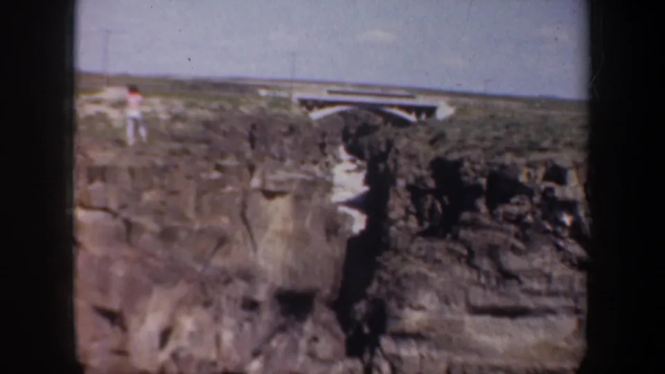 A distant visitor stands above the rocky canyon, with a low bridge and open landscape visible beyond the rim.