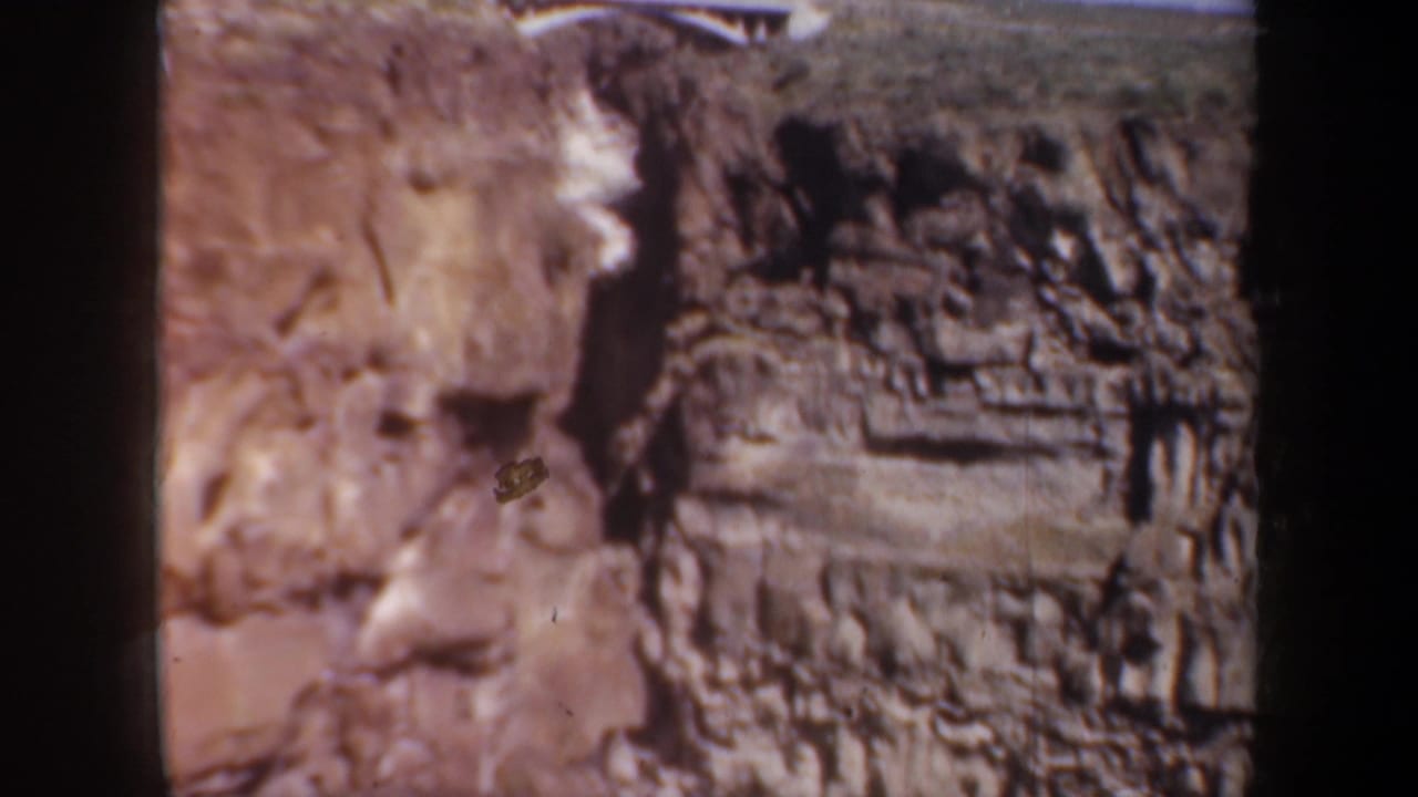 1957 Oregon home-movie frame showing a sunlit layered rock wall and a small white fence near the upper edge.