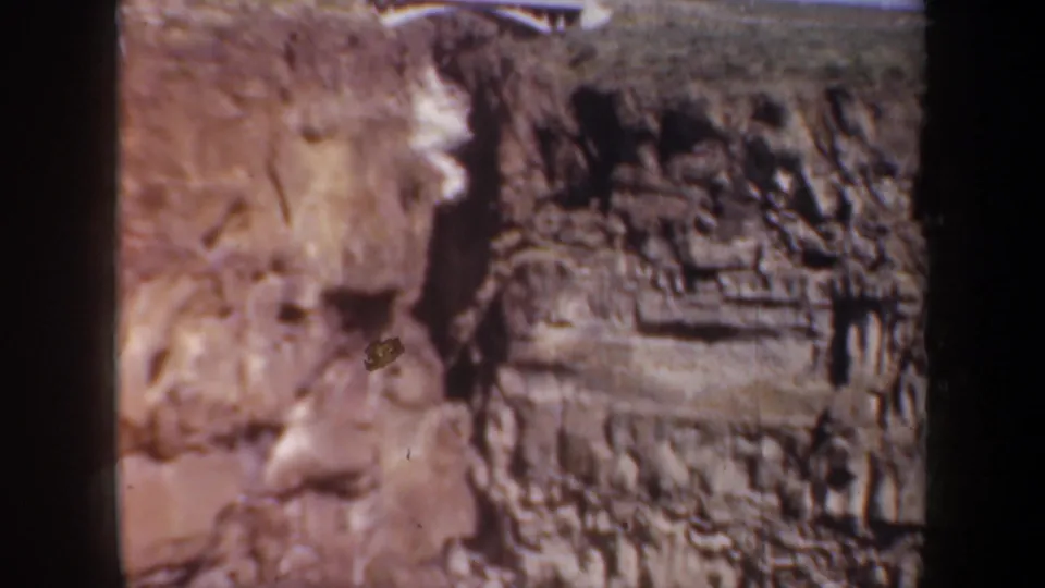 1957 Oregon home-movie frame showing a sunlit layered rock wall and a small white fence near the upper edge.