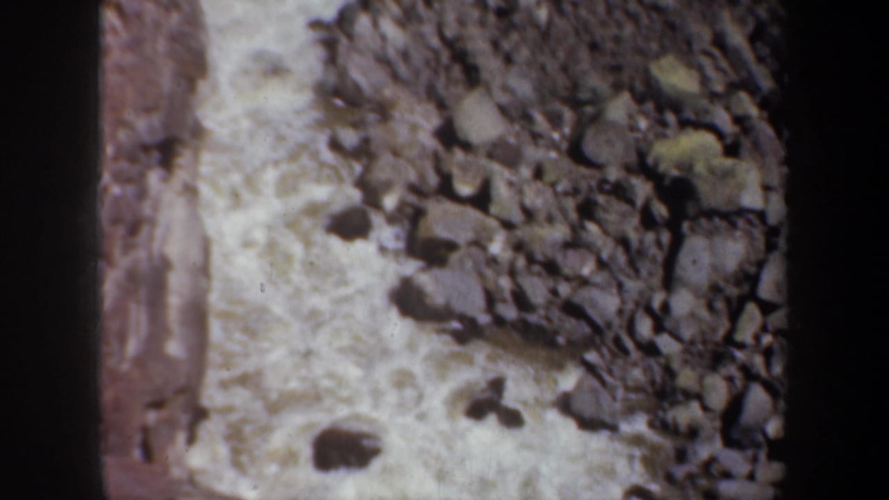 1957 8mm footage shows churning water running along a rough bed of stones, framed tightly with dark film edging.