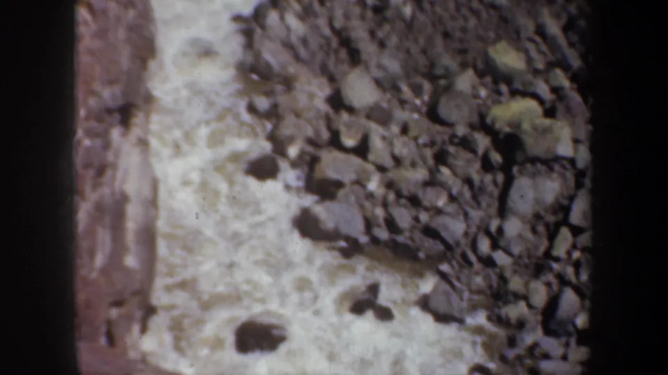 1957 8mm footage shows churning water running along a rough bed of stones, framed tightly with dark film edging.