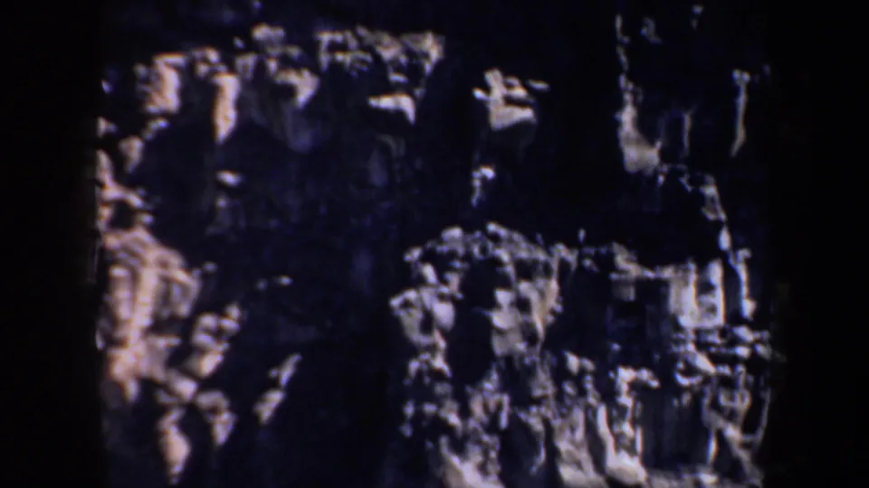1957 8mm view of a steep, shadowed rock formation with pale ledges and textured cliff surfaces filling the frame.