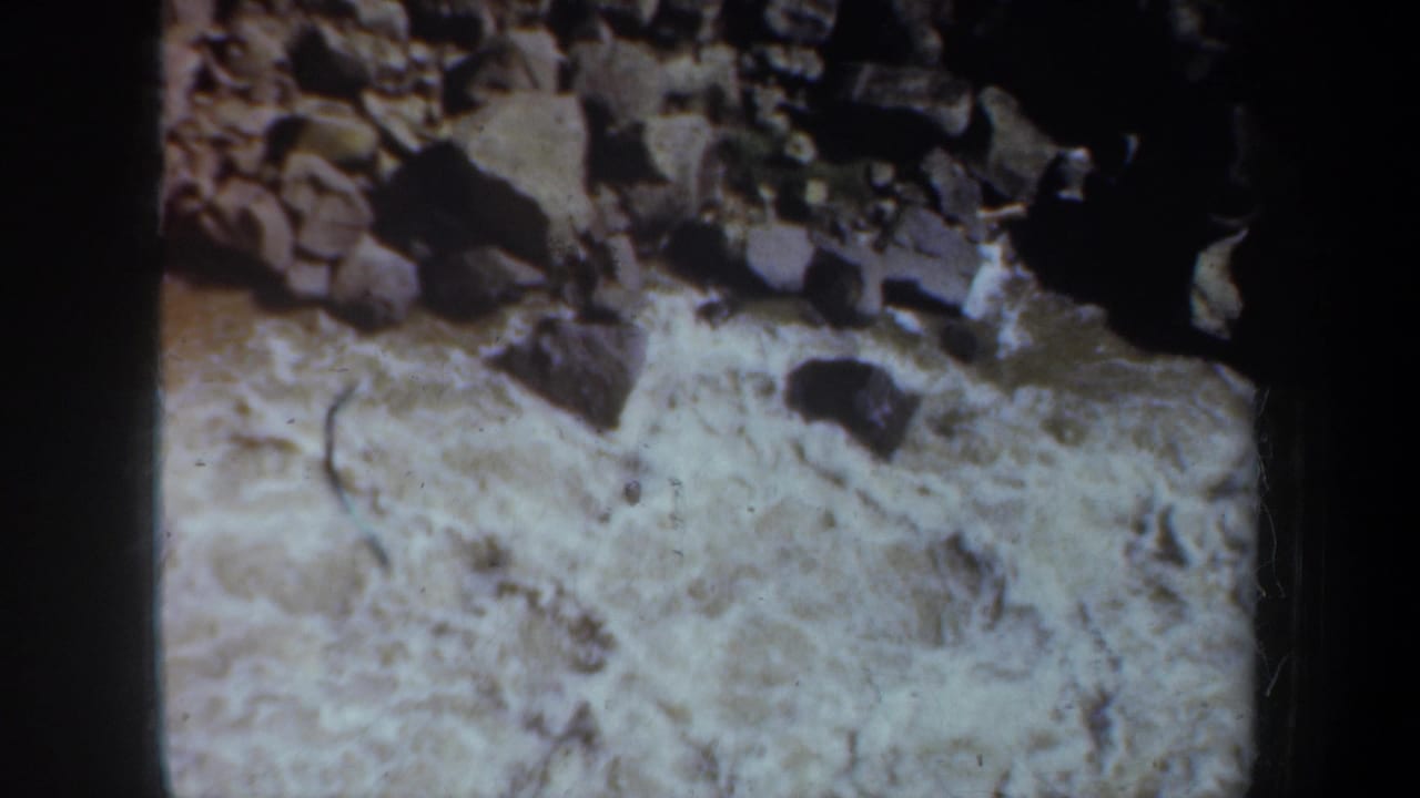 Close 8mm view of pale flowing water and dark river rocks in the Oregon-associated home-movie landscape sequence.