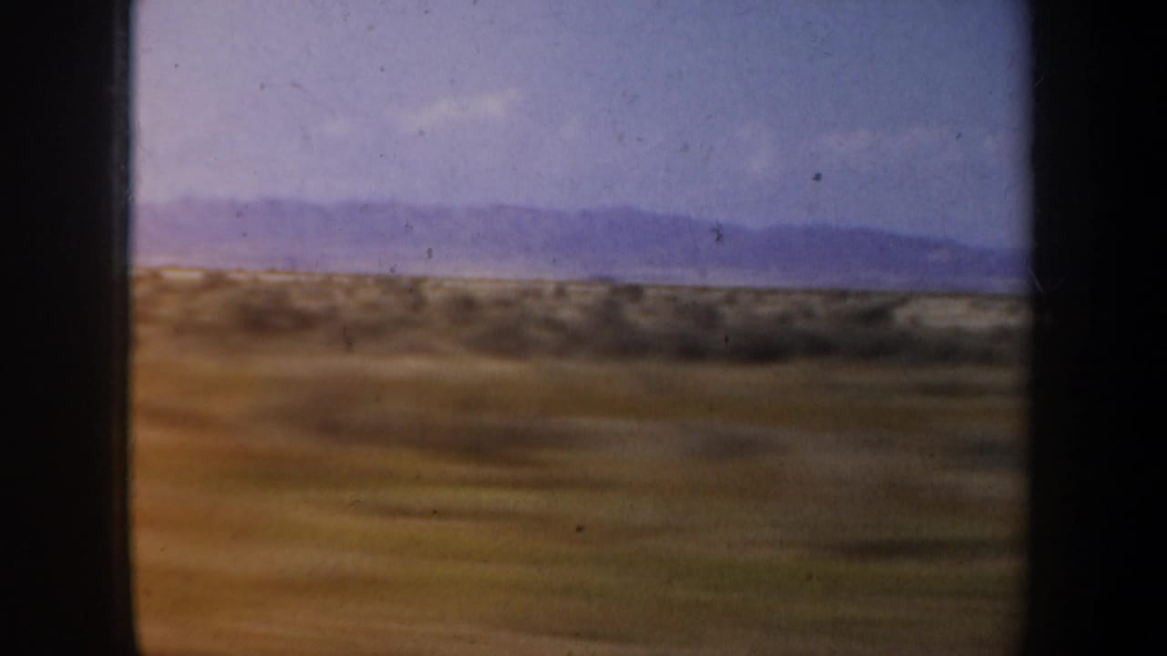 Blurred scrubland and low hills passing below a pale sky and distant mountains.