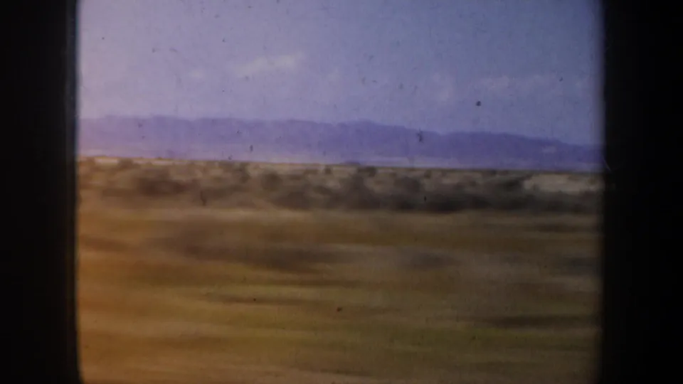 Moving 8mm road-view frame with blurred brown scrubland, low hills, pale lavender sky, and a far mountain ridge.