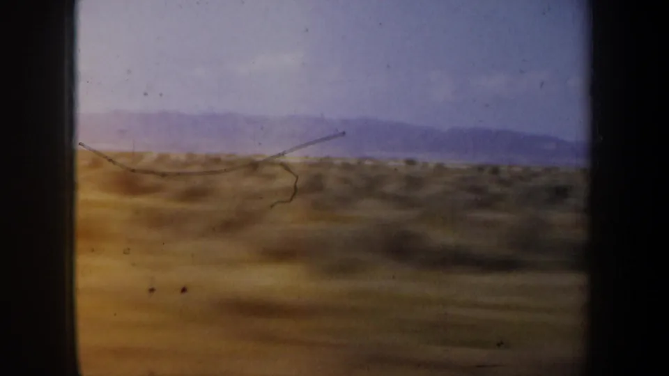 1957 8mm frame of brown roadside terrain and far mountains, with visible scratches and motion blur from travel.