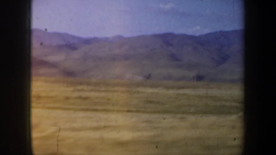 Cataloged 1957 8mm travel footage showing dry open terrain and distant mountain ridges through a dark vehicle window edge.