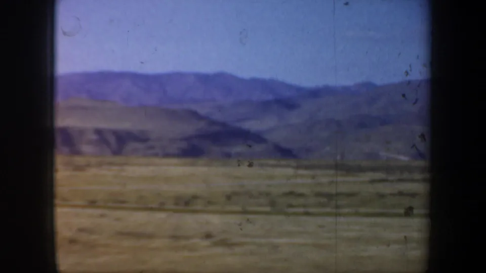 Soft 8mm frame of a purple-toned mountain ridge and broad scrubland, viewed from inside a moving vehicle.