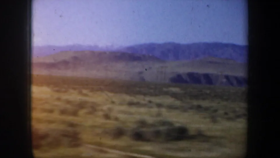 Cataloged 1957 home-movie frame showing arid foreground, low hills, and distant mountains through dark vehicle-window framing.