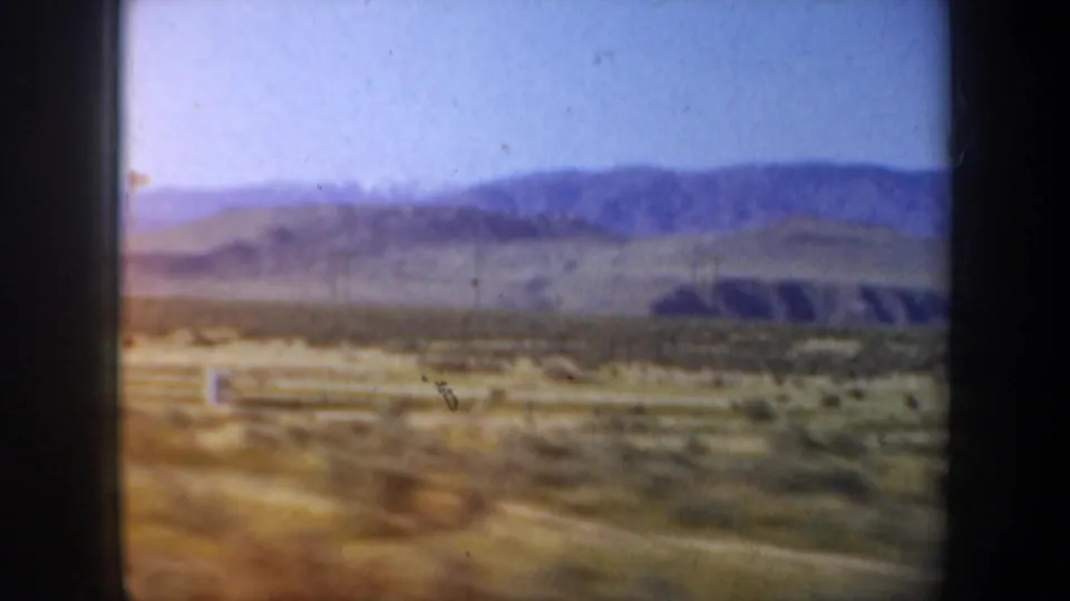 Soft 8mm travel frame with a white fence or roadside barrier, scattered trees, and distant mountain ridges in the background.