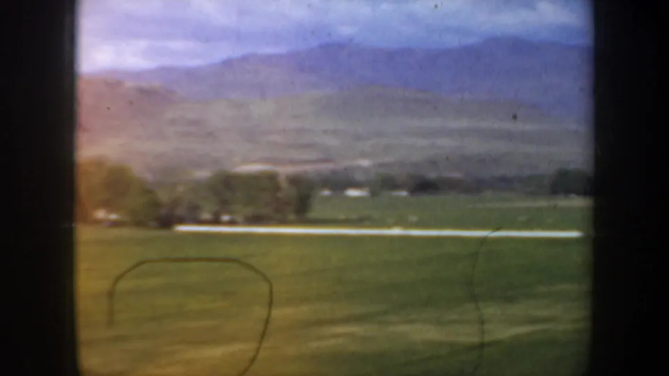 Wide 1957 rural Oregon view with open green acreage, scattered trees, a pale road or fence line, and layered hills beyond.