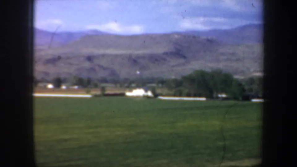 Rural Portland area 8mm frame showing pasture, small distant farm buildings, a tree cluster, and low mountains beneath a cloudy blue sky.