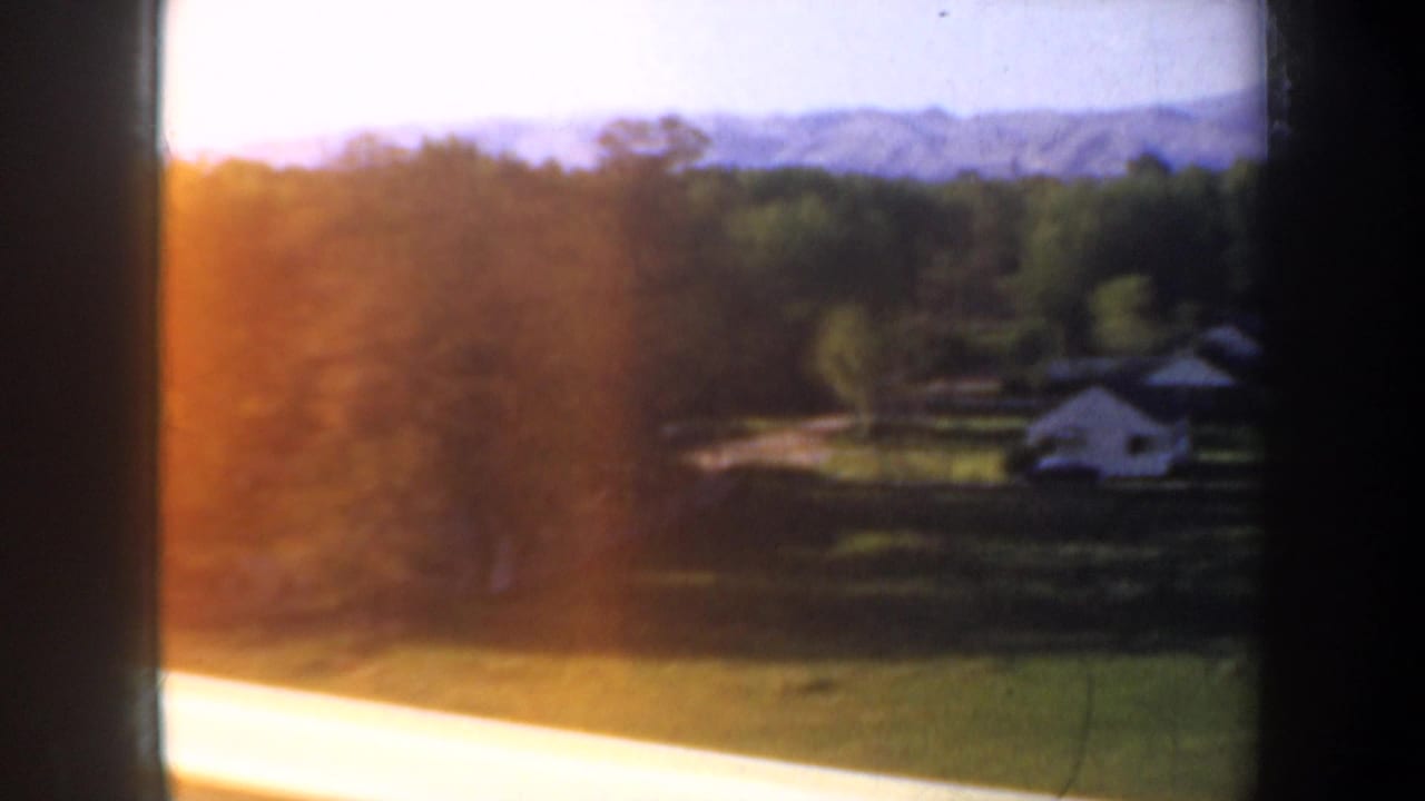 Bright countryside view with field, houses, trees, hills, and strong sunlight at the window edge.
