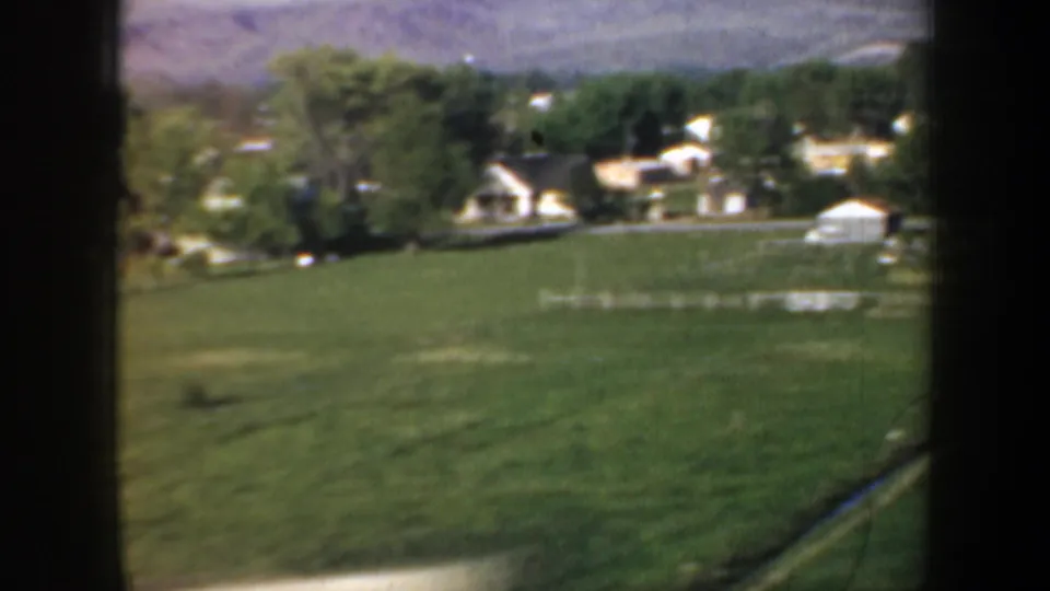1957 Portland-area countryside seen from a moving vehicle, with a broad field, clustered houses, trees, and low hills beyond.