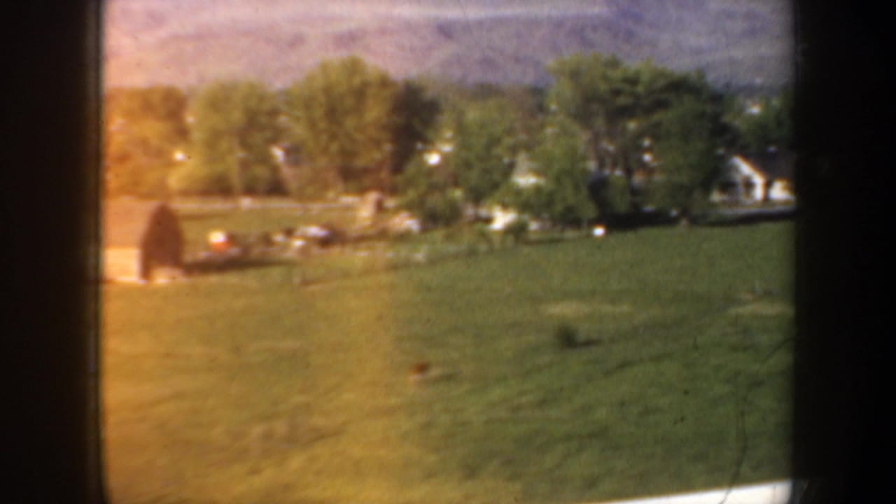 1957 Portland-area home-movie view of a green pasture, trees, and small light buildings through a vehicle window.
