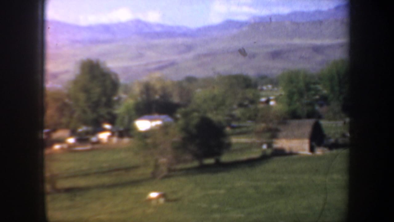 Rural field, scattered buildings, trees, and distant hills under a blue sky.
