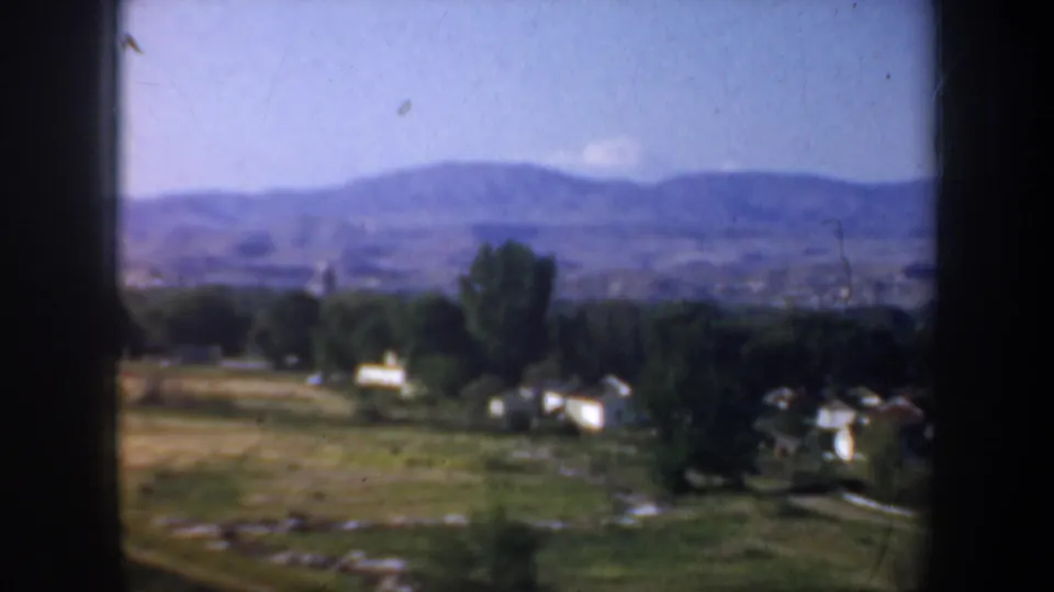 1957 8mm view of fields, trees, and small white buildings with hazy hills beyond the Portland-area landscape.