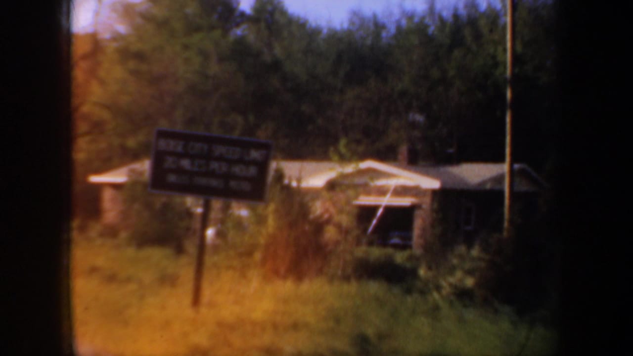 1957 Portland-area home-movie frame showing a roadside house partly hidden by foliage with a dark sign in front.