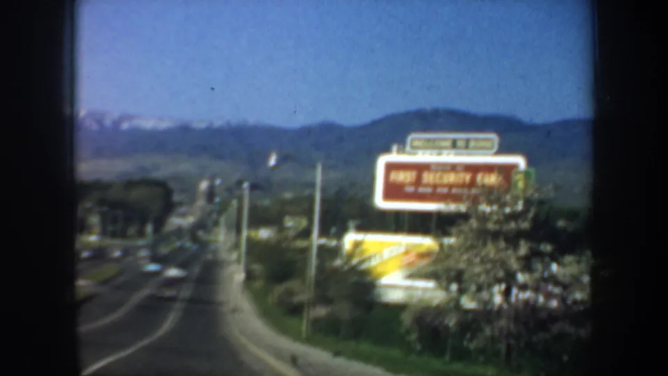 A moving-vehicle view shows a highway with cars and a prominent First Security Bank roadside billboard below mountain foothills.