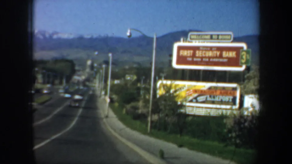A highway scene shows First Security Bank and Allpost signage near passing cars, utility poles, green hills, and distant mountains.