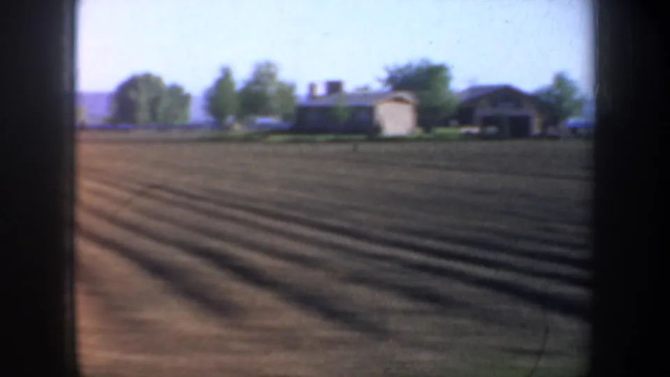 1957 Oregon roadside frame showing cultivated rows, distant homes, trees, and low hills from a moving-car perspective.