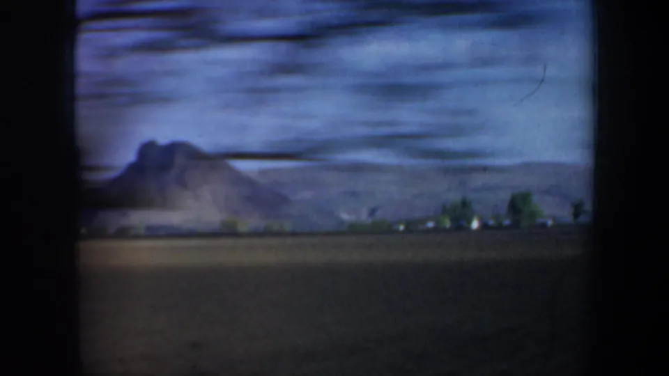 Motion-blurred 1957 Oregon home-movie frame showing open farmland, a distant hill or mountain, and small structures at its base.