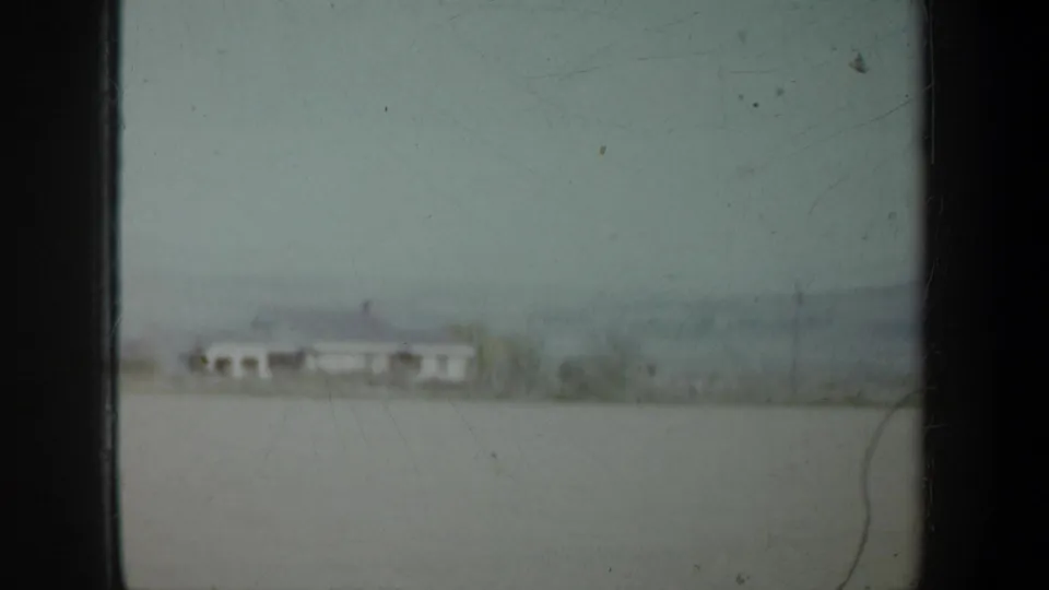 Faded 1957 Oregon home-movie view of a modest rural house set in open land with low hills or haze in the background.